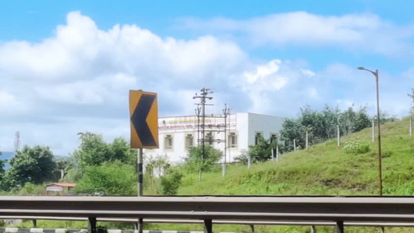 An Indian flatbed truck with an empty, open trailer travels on a winding highway under a partly cloudy sky. Visible road signage indicates a turn, confirming a South Asian setting with elevated terrai