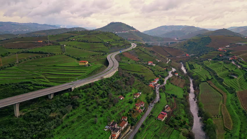 Stunning countryside greenery aerial view. Curve asphalt road running vivid slopes under cloudy sky. Small stream crossing lush green fields with village buildings. Picturesque gloomy rural landscape.