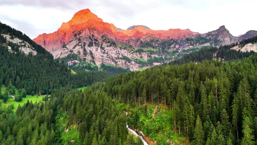 Sunrise illuminates alpine peaks above Lenk Simmenfälle, Switzerland, with vibrant orange glow, dense pine forest stretching across the valley, and a waterfall flowing through the lush green landscape