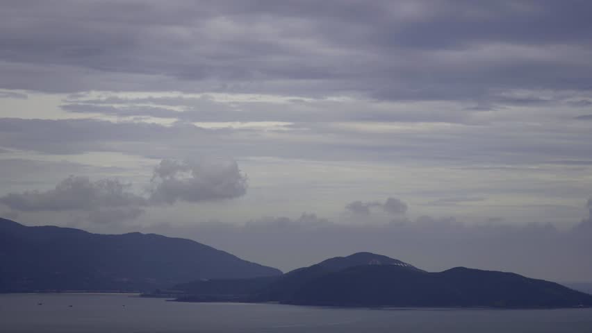Panoramic view of a serene coastal landscape featuring dark, rolling mountains meeting tranquil bay waters under a vast, dramatic overcast sky with soft clouds.