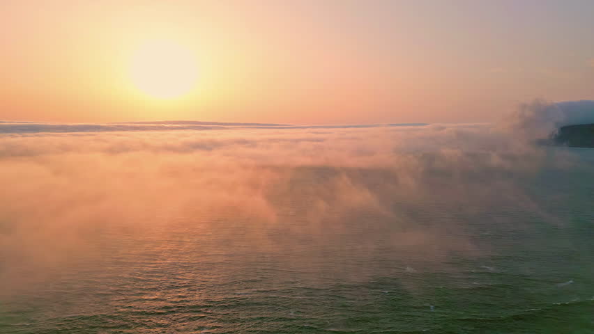 Amazing ocean sunset illuminating white fog covering calm water aerial view. Golden sun disc setting over sea horizon with soft evening mist. Picturesque summer evening drone shot. Waves rippling