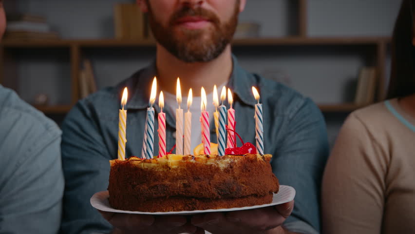 Close up unrecognizable man holding birthday cake with burning candles happy male smiling make wish near friends three unknown people at home festive party celebrating event together congratulation