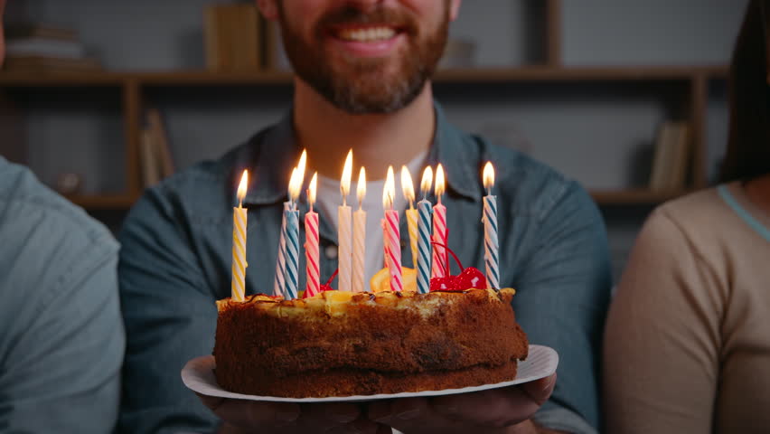 Close up unrecognizable man holding birthday cake with burning candles happy male smiling make wish near friends three unknown people at home festive party celebrating event together congratulation