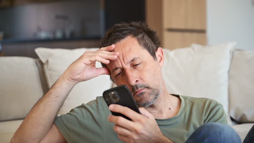 Tired lazy man alone at home sitting on sofa in casual clothes holding smartphone ad yawning. 4K with selective focus