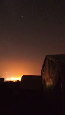 Star trails sweep above Mount Ararat base camp tents, orange city glow on horizon, rocky ridge silhouettes under dark sky during long-exposure night time lapse.