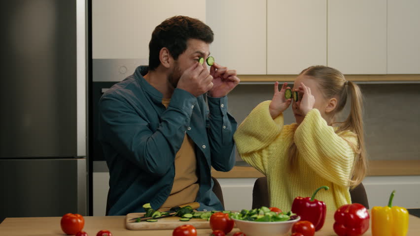 Happy Caucasian family at kitchen playing fooling having fun Caucasian child girl daughter kid with father holding cucumber slices like glasses cover eyes with vegetable circles joyful funny cooking