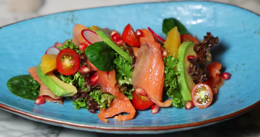Vibrant gourmet salad with smoked salmon, avocados, cherry tomatoes, pomegranate seeds on blue plate. Fresh, healthy dining concept at upscale restaurant, top view.