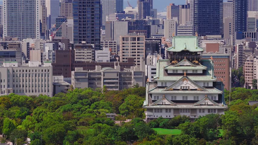Slow sweeping aerial view of Osaka Castle surrounded by green trees, transitioning to modern skyscrapers and the urban landscape of Osaka, Japan.