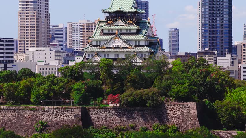 Aerial view of Osaka Castle with its green roof, surrounded by greenery, panning to reveal the urban skyline of Osaka, Japan.