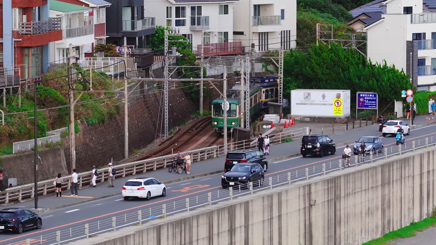 Aerial view of a green and cream train moving along tracks beside a busy road in Kamakura, Japan. Cars, bicycles, and pedestrians add to the urban scene.