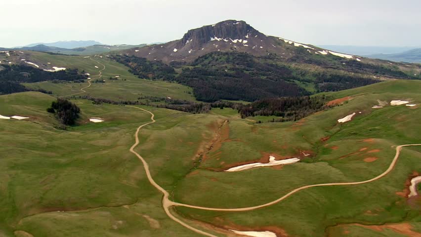 A winding dirt road leads through green hills towards a majestic mountain