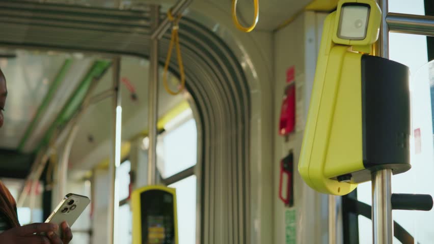 Charming African American woman holding smartphone close to yellow validator paying for ride on bus. Passenger in striped shirt and jacket completing contactless payment. Modern technology. - Powered by Shutterstock - Get 15% off with code: PIKWIZARD15