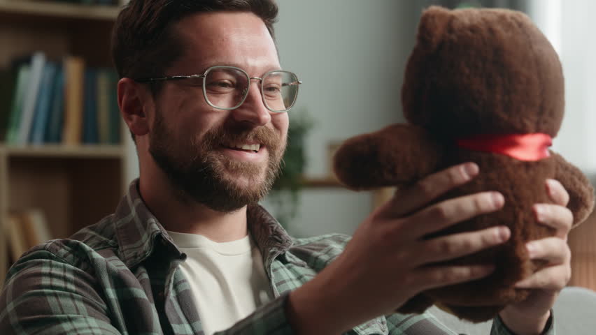 Adult happy Caucasian man indoors holding fluffy toy Teddy Bear preparing gift present for child kid middle-aged carefree male at home positive guy sitting on couch in living room homeowner father