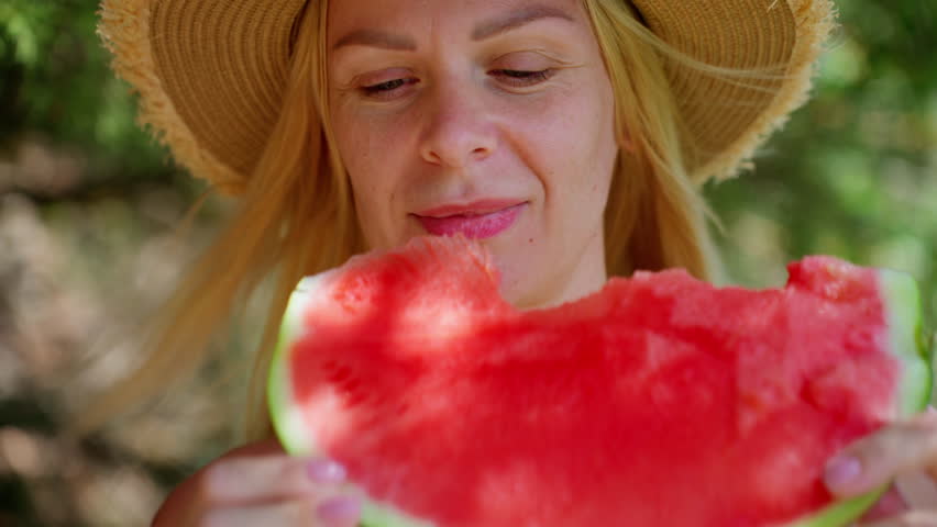 Beautiful blonde woman in straw hat enjoy fresh, juicy slice of watermelon on sunny day. Healthy summer lifestyle concept with happy person eating ripe fruit in nature
