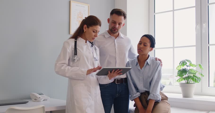 Female family doctor in medical office showing examination results on a tablet screen and having consultation with young married couple planning pregnancy standing in clinic. 4k video.