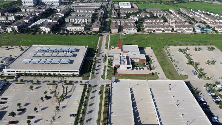 Large surface parking lot, rooftop ramp garage separated by grassy land from townhomes and offices, infrastructure layering, suburban planning, and future development potential in North Texas. USA