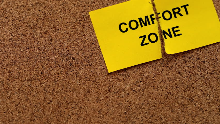A ripped yellow paper note with the words Comfort Zone on it lying on a cork board. Close up.