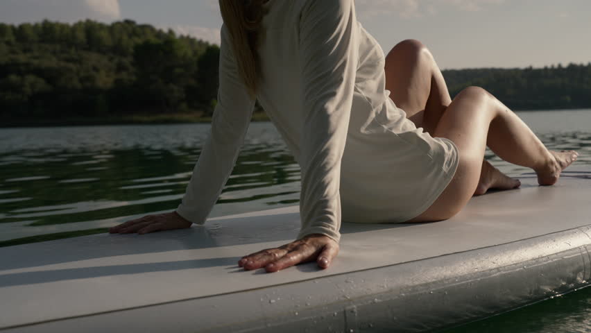 Blonde woman in white athletic bodysuit and cap sits on paddleboard, soaking up sun on calm lake, enjoy peaceful nature, summer relaxation and wellness getaway