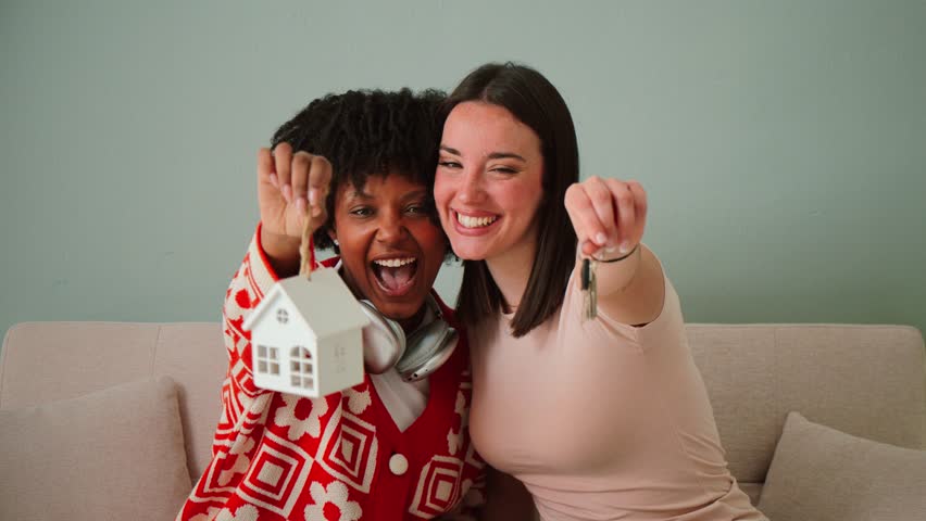 Two ecstatic females proudly displaying a house key and a tiny house model, celebrating their new home ownership journey with immense joy and enthusiasm, radiating happiness and excitement in a warm