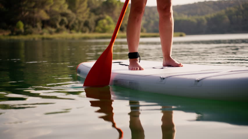 Close-up view of feet on stand up paddle board as person paddles on calm lake, with beautiful reflections on water surface during serene summer day creating peaceful atmosphere
