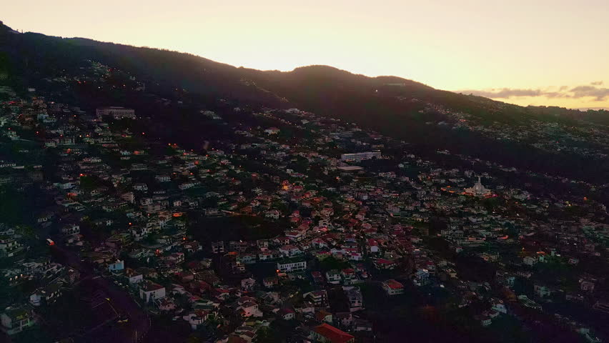 Bright sunrise shining behind hills covered with cozy town streets. Orange morning sun rising on horizon. Beautiful small settlement stretching on green slope to yellow skyline. Tiled roof houses.