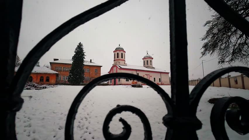 Winter scene with a snow-covered church and school in a rural village. Captured with smooth handheld panning through a blurred metal fence in the foreground during snowfall.