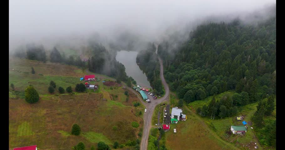 Aerial 4K view of Balıklıgöl Plateau in Trabzon, Turkey, with misty mountains, a forest-lined lake, scattered houses, and lush greenery creating a tranquil and scenic highland atmosphere.