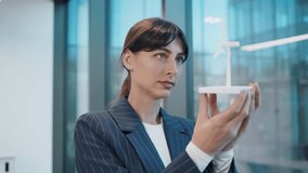 Close up portrait of elegant Caucasian businesswoman examining small wind turbine model in hands. Female professional analyzing clean energy prototype in modern office near window. Indoors. - Powered by Shutterstock - Get 15% off with code: PIKWIZARD15