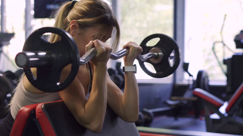 Upper Body Strength: Blonde woman executing concentrated bicep curls with a bar at the gym