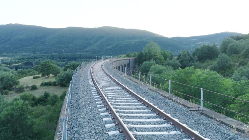 Empty train tracks curve through a verdant valley, disappearing into the distance under a soft, cloudy sky