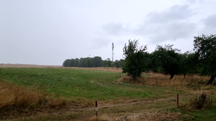 Strong wind and heavy rain hitting a green field with trees and a telecommunication tower