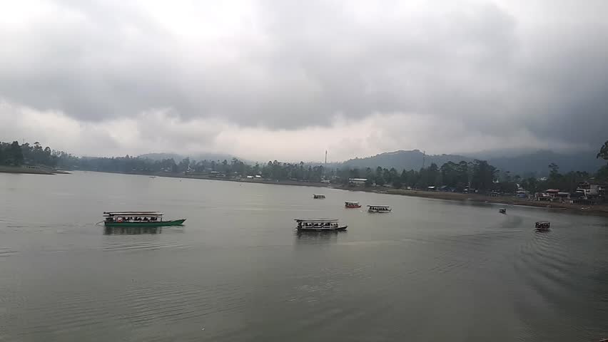 Enjoying the natural scenery and the tranquility of the lake on a boat is a fun recreational activity, the sky looks cloudy, the land is filled with trees and hills, making for refreshing backdrop.