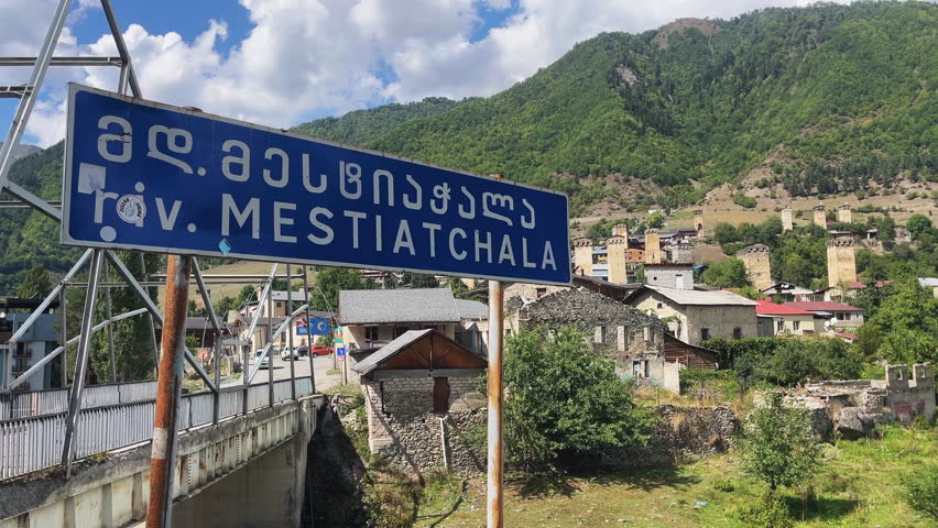 View of a blue road sign in Georgian marking Mestiatchala, with cars crossing the bridge, the village of Mestia behind, medieval defense towers, and mountains in the background.