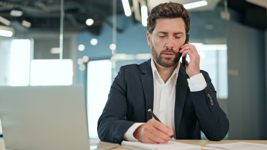 Businessman makes important phone call at modern office desk. Dressed in dark suit successful male writes notes expressing focus productivity. Cheerful man enjoys conversation in business environment.