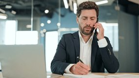 Businessman makes important phone call at modern office desk. Dressed in dark suit successful male writes notes expressing focus productivity. Cheerful man enjoys conversation in business environment. - Powered by Shutterstock - Get 15% off with code: PIKWIZARD15
