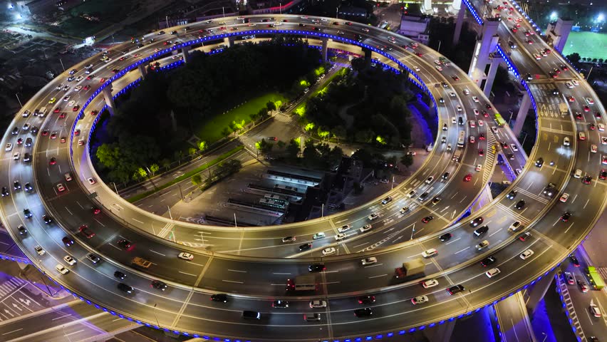 Aerial view of a spiraling highway interchange with heavy vehicle traffic and blue illumination at night in Shanghai, China.
