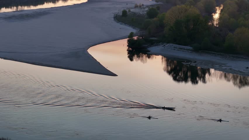 drone shot captures rowing boats leaving glittery trails on the River Po at sunset between Lombardy and Emilia, with golden light reflecting on the water and a wide sandbank visible, slow motion
