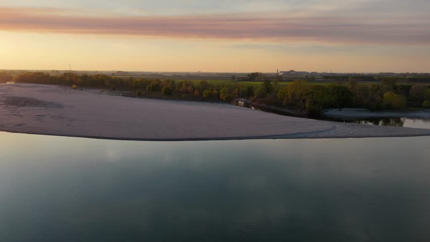 drone shot captures the River Po flowing at sunset, with the Lombardy countryside stretching across the water from Emilia Romagna, fields and trees glowing under a soft sky in Piacenza, slow motion