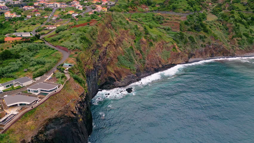 Small town on coastal cliffs aerial view. Cozy settlement standing on high green rock washed by blue ocean water. Picturesque waves crashing on rugged stone shoreline. Beautiful sea splashing on coast