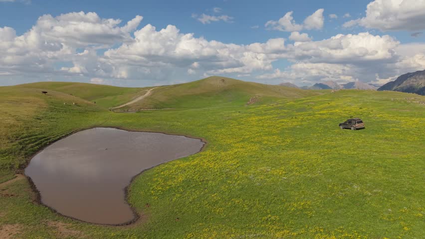 Green meadow with yellow flowers and a small pond reflecting the clouds, an suv is parked near the pond, with mountains and hills in the background