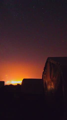  Star trails sweep above Mount Ararat base camp tents, orange city glow on horizon, rocky ridge silhouettes under dark sky during long-exposure night time lapse.