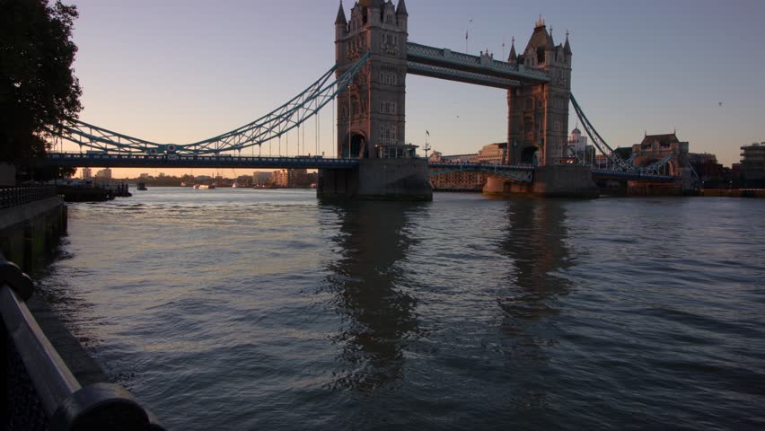 Slow motion sunrise illuminates Tower Bridge’s stone towers and blue suspension, calm Thames water mirrors golden sky, serene wide view blends city’s history and early morning glow