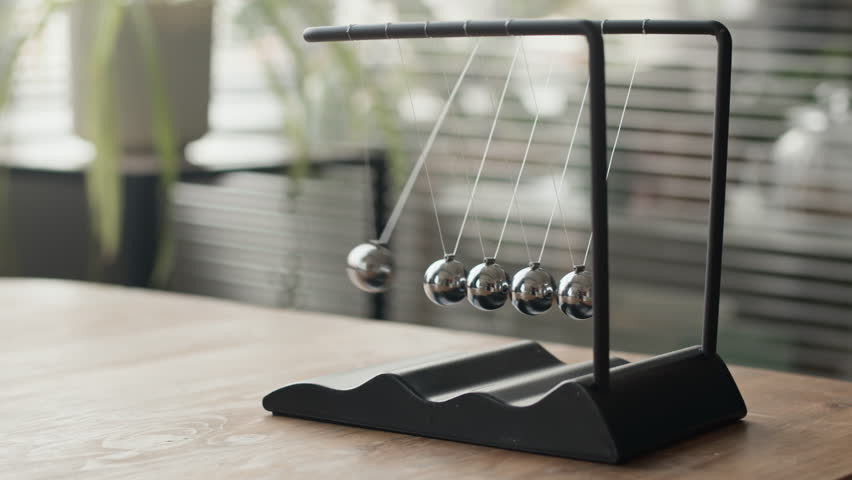 Closeup no people shot of Newtons cradle on office table with one steel ball striking spheres and transmitting pressure wave, demonstrating principle of conservation of energy