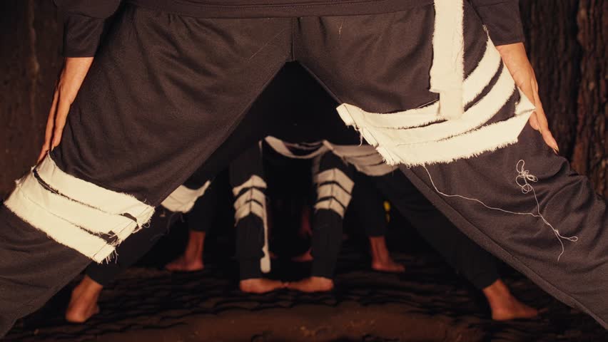 A troupe of barefoot dancers in dark, contemporary costumes performs a synchronized, rhythmic movement in a mysterious passage. This experimental piece is viewed from a unique low angle.