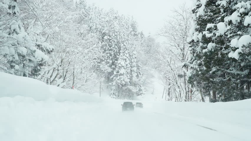 Point of view by windshield of Driving car on a snow-covered mountain road with pine tree forest in snowy day. Beautiful scenic nature countryside and transportation in winter season.