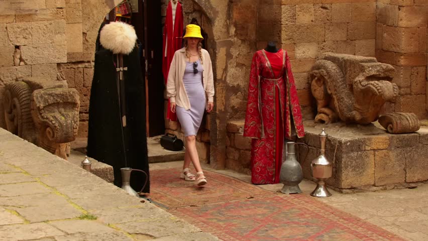 Young Caucasian woman in a yellow hat walks through a historic marketplace. Traditional clothing and artifacts are displayed nearby, creating a vibrant cultural scene.