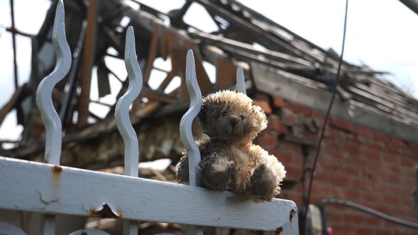 Dirty teddy bear sits on fence symbolizing lost childhood and broken peace with blur destroyed house at background. Scene reflects pain and tragedy caused by russian invasion and violence in Ukraine