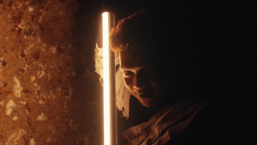 In a dark, gritty tunnel, a man with white contact lenses is illuminated by a vertical fluorescent light. He stares with a disturbed and manic expression, full of fear and intensity.