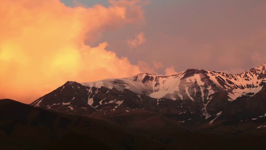 Snow-capped mountains under a colorful sunset sky. The landscape features rugged peaks and a dramatic cloud formation.