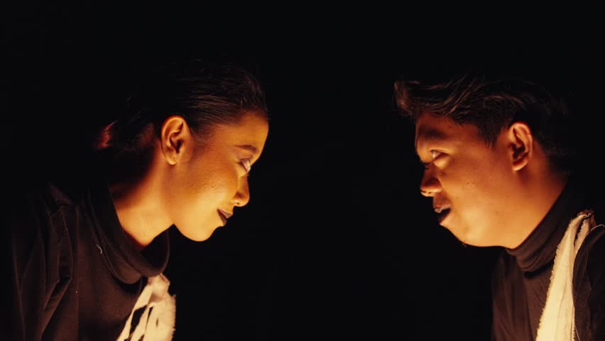 Profile view of a man and woman with dramatic makeup staring intently at each other in the dark. Lit from below, their serious expressions create a scene of tense confrontation or intimacy.